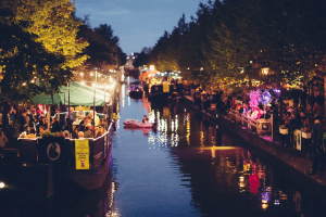 A nighttime picture of a brightly lit canal in the Netherlands, where people are enjoying themselves on boats and along the canal banks, reflecting a festive evening atmosphere.