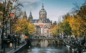 A scenic view of a canal in Amsterdam with a large church in the background and autumn trees on the banks, showing the beauty of Holland.