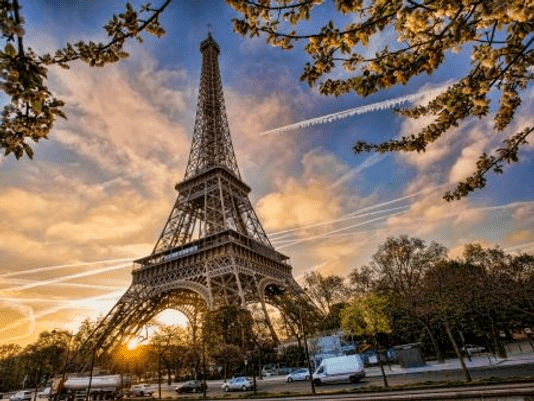 The Eiffel Tower in Paris, framed by blossoming tree branches, stands majestically against a vibrant sky with streaks of clouds at either sunrise or sunset, while traffic moves along a street below.