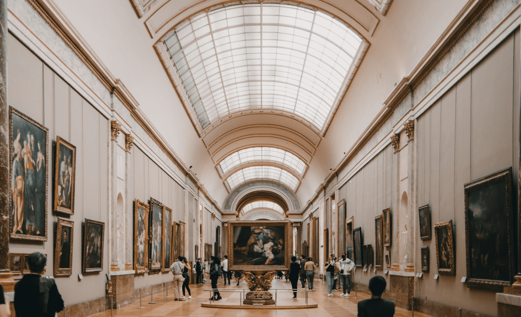 An interior shot of a long, elegant hall in the Louvre Museum in Paris, with a vaulted glass ceiling, where people are admiring numerous large paintings on the walls.