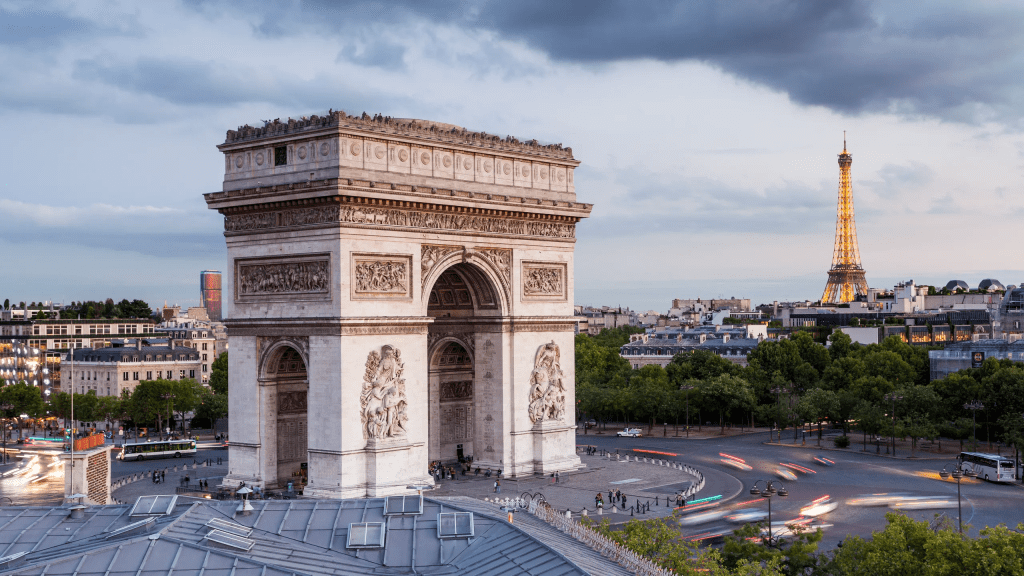 An outdoor shot of the Arc de Triomphe in Paris, a large neoclassical monument with detailed bas-relief sculptures on its facade, set against a bright blue sky with a few clouds.