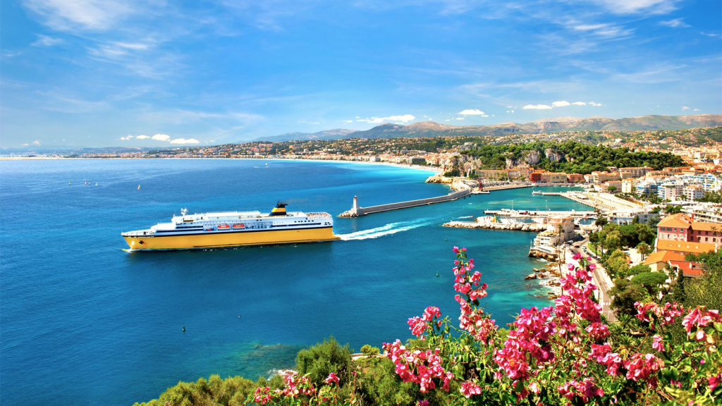 Riviera coastline with a large yellow ferry leaving a harbor in a coastal city. The beautiful blue sea is in the foreground, with pink flowers and lush green hills under a bright, partly cloudy sky.