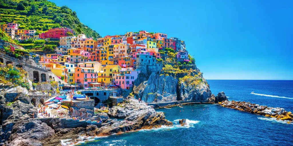 Riviera village with brightly colored houses built on a steep cliffside overlooking the deep blue sea. The picturesque coastal scene features rocky cliffs and a small harbor on a sunny day.
