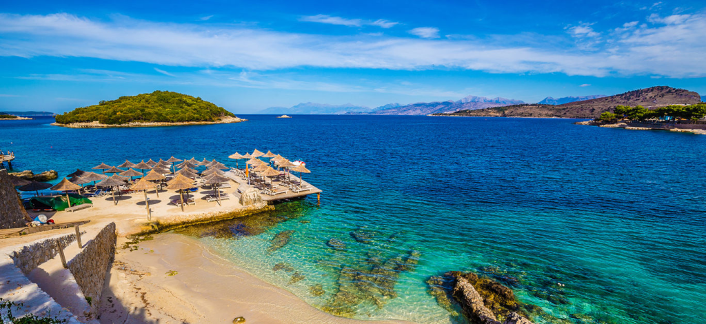 Riviera beach with a small pier, straw umbrellas, and lounge chairs on a shoreline of turquoise water. A lush green island and mountains are visible in the distance under a bright blue sky.