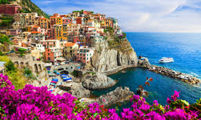  village with brightly colored houses built on a steep cliffside overlooking the deep blue sea. The picturesque coastal scene is framed by vibrant pink flowers in the foreground on a sunny day.