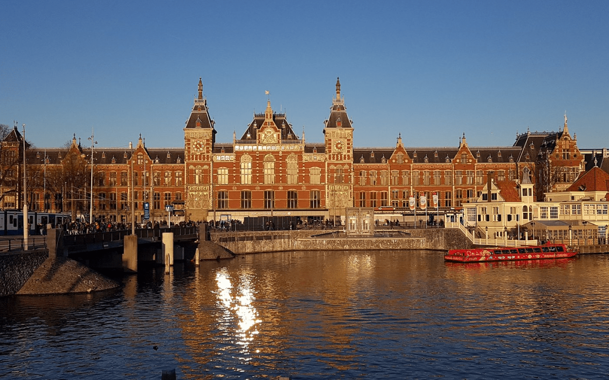 A picture of Amsterdam Central Station, a prominent landmark in tourism in the Netherlands, showing its historic building overlooking the city's canal on a sunny day.