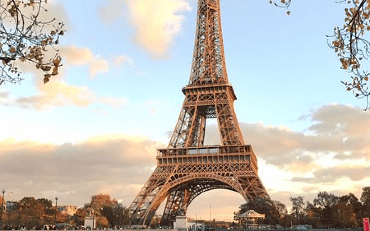 The Eiffel Tower in Paris stands tall against a partly cloudy sky, viewed from across the Seine River with a bridge in the foreground.
