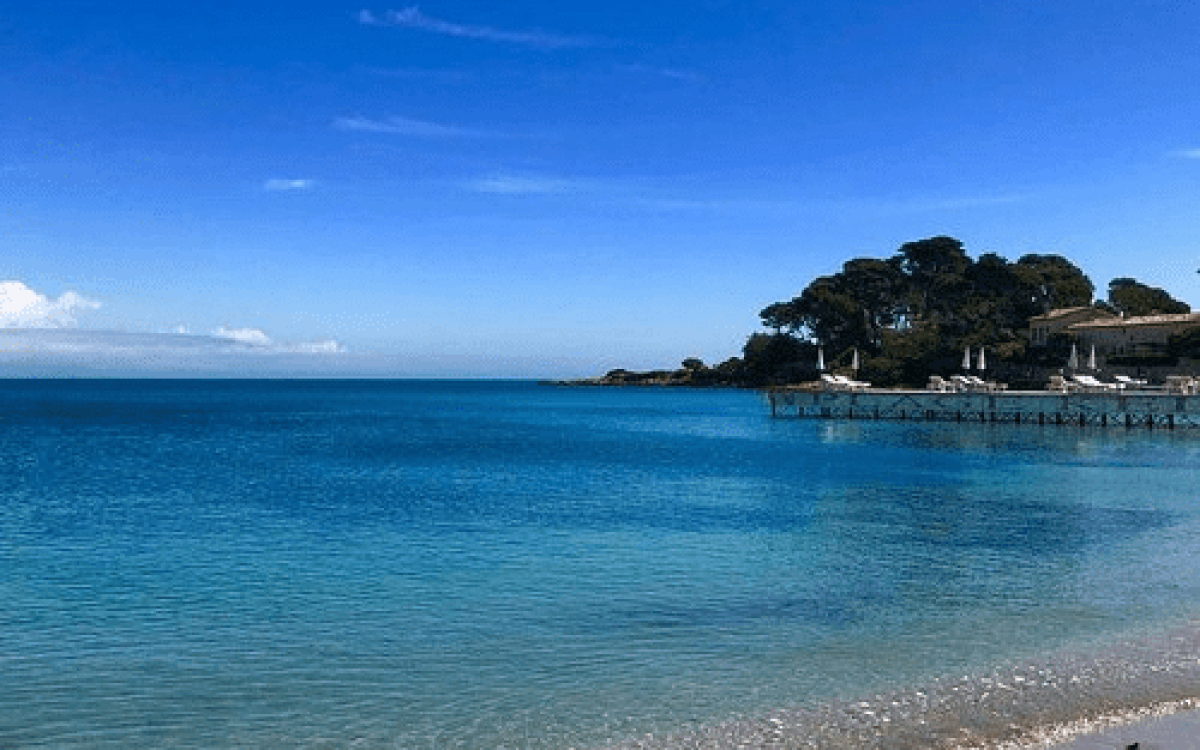 Riviera beach with clear turquoise water, a sandy shore, and a lush green headland with a pier and buildings in the background under a bright blue sky.