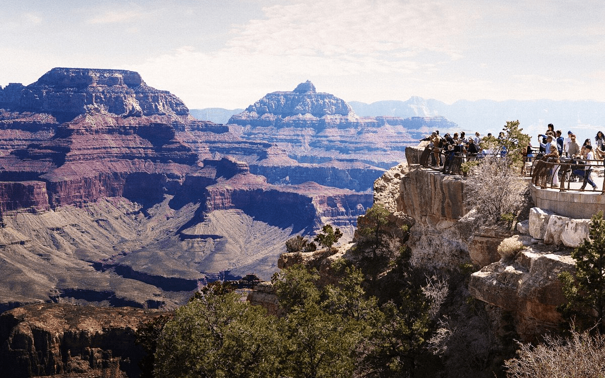 A large group of tourists stands on a viewing platform, admiring the vast, multi-layered rock formations of the Grand Canyon. The scene captures the immense scale of the canyon under a bright sky with scattered clouds.